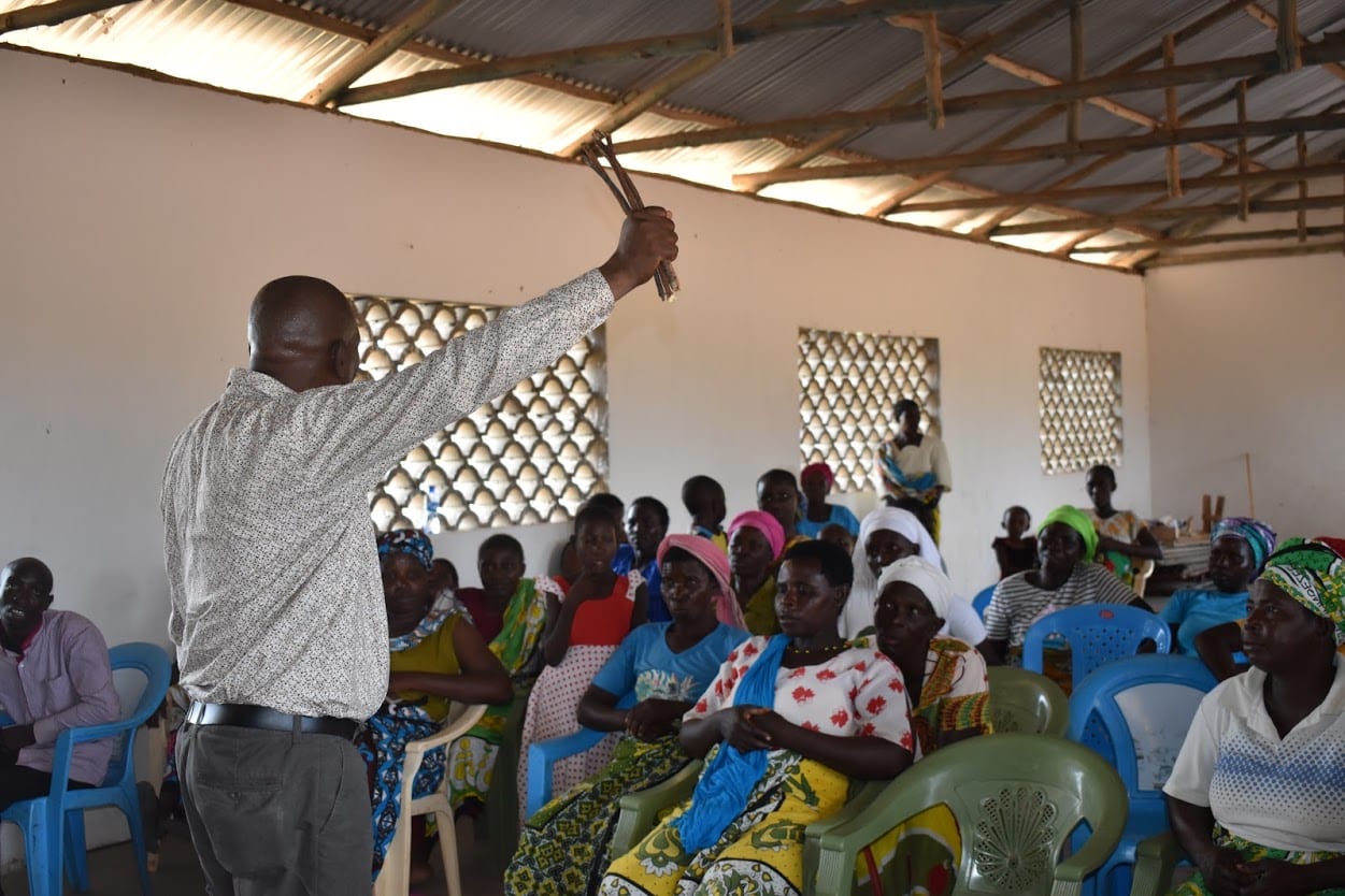 A Kenyan man standing before a group of 20 listeners with one arm raised.