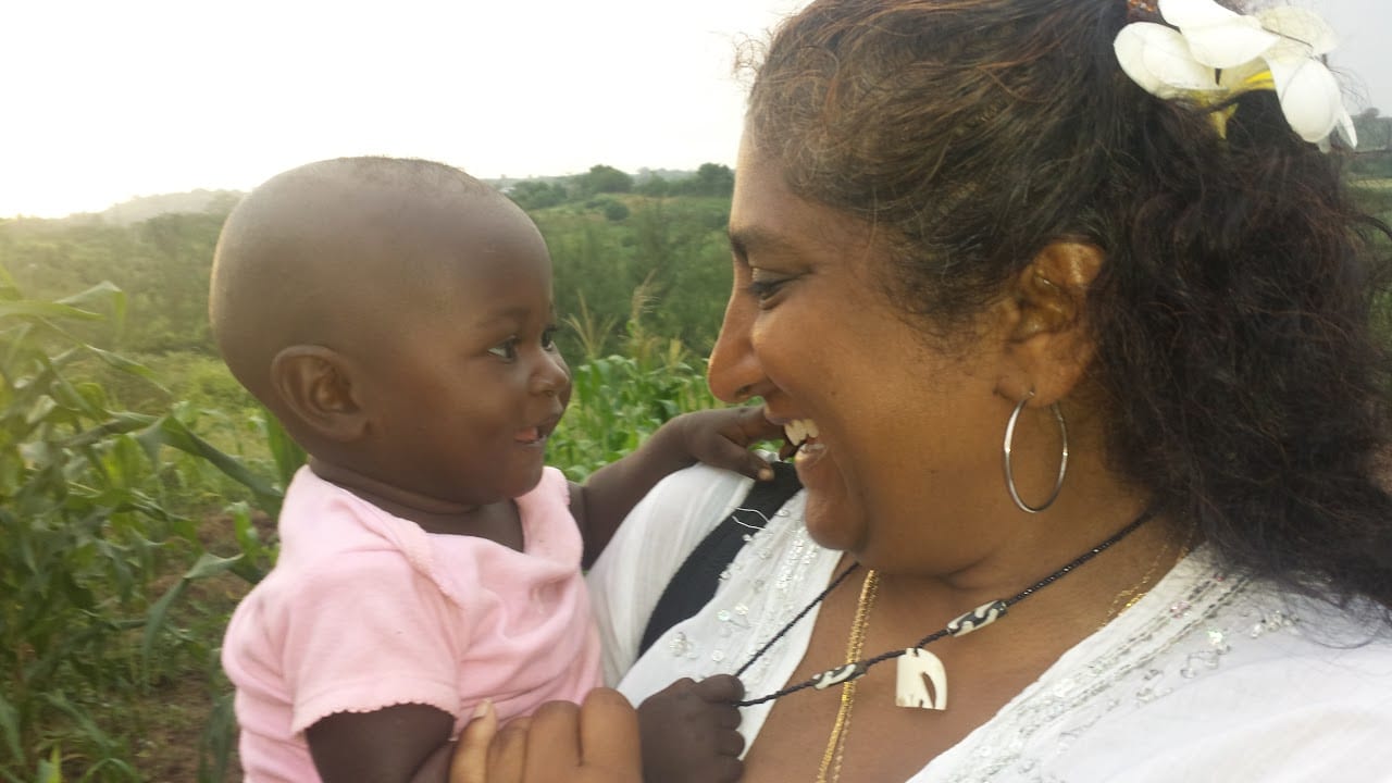 An adult woman standing outside, smiling a baby girl that she is holding.