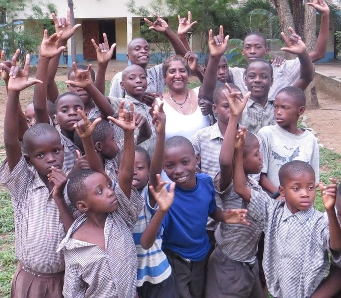 A woman standing in the middle a group of smiling young boys all holding up the sign-language gesture for "I love you".