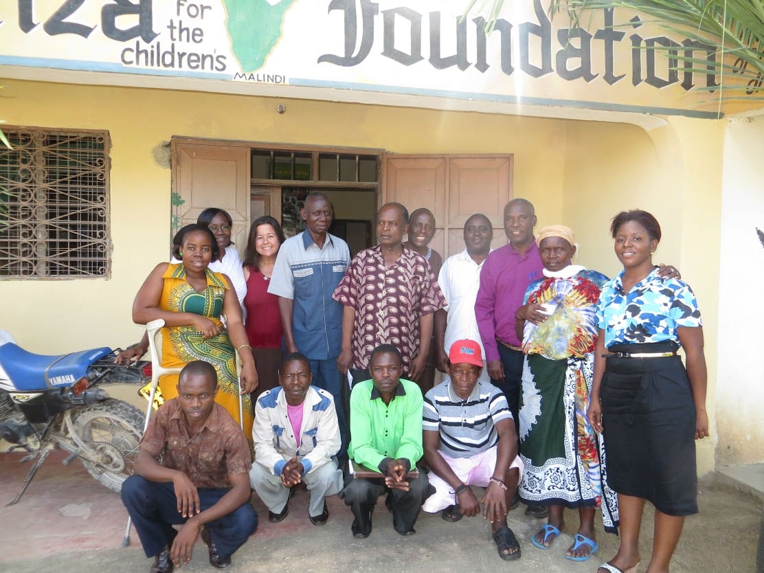 14 adults standing outside of a building, smiling at the camera