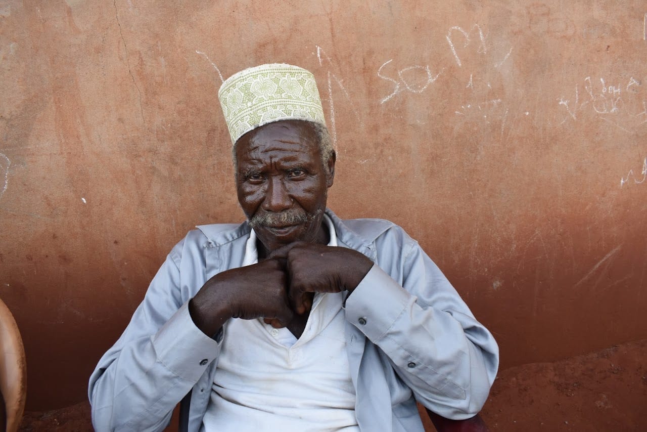 An elderly man sitting before a red wall wearing patterned hat.