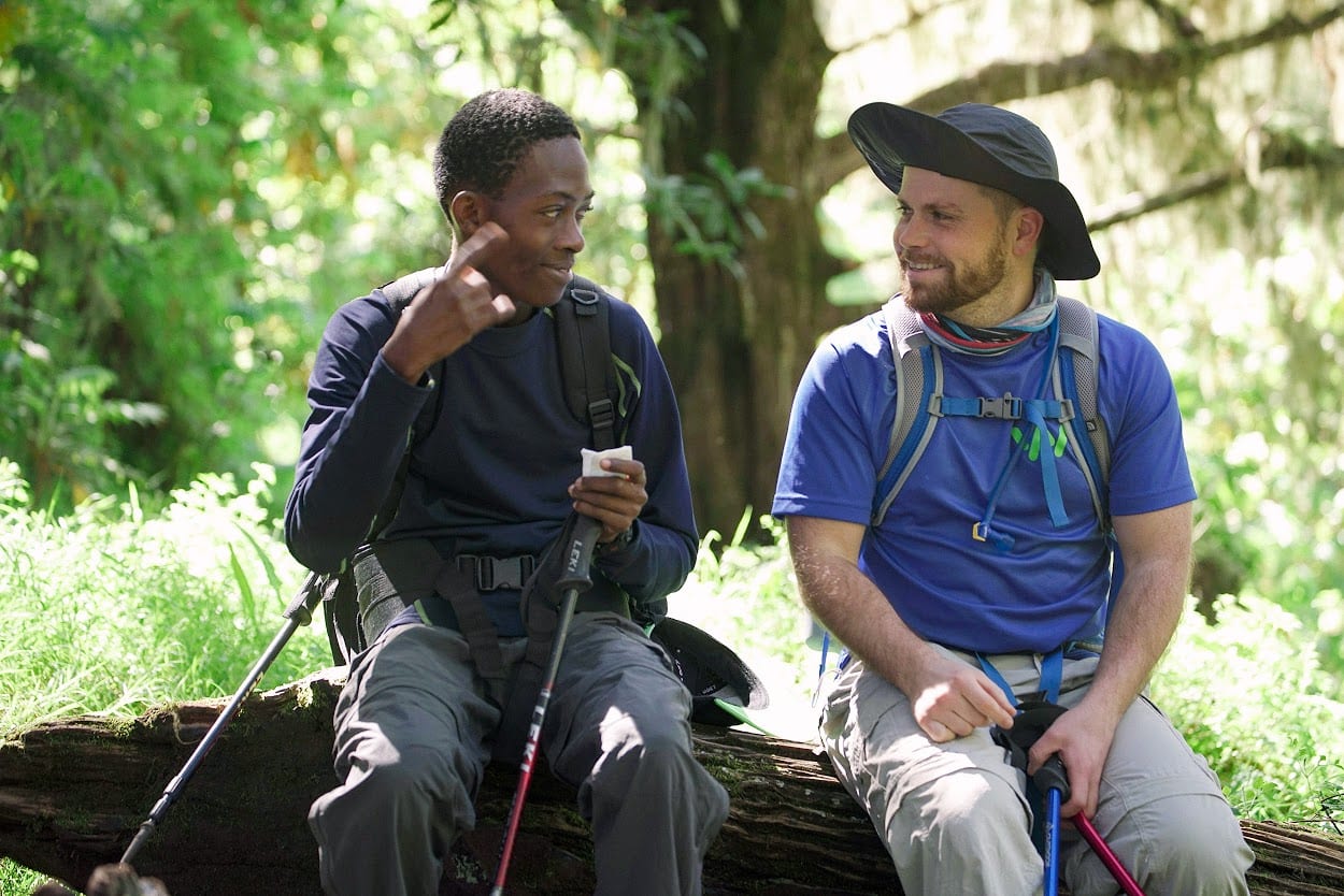 Two young mean seated side by side on a log, smiling at each other.