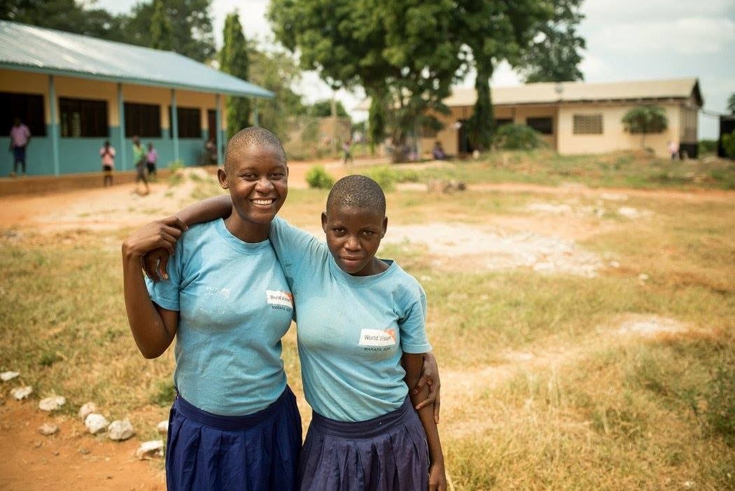 Two Kenyan girls standing outside their school with their arms around each other.