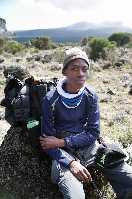 A teenage male seated on a rock overlooking a mountain, has a hiking backpack behind him.
