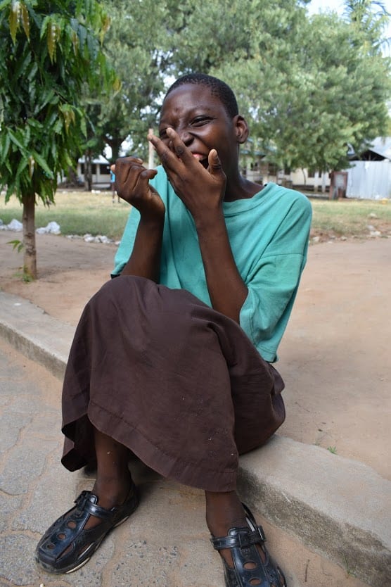 A young girl wearing a blue shirt, sitting on the ground outside, smilig with her hands in front of her face.