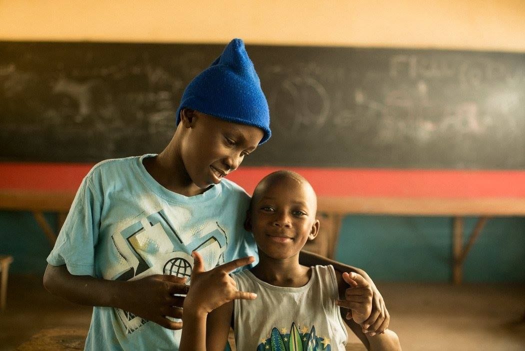 Two young boys standing side by side, one is wearing a blue hat the other is making a hand gesture.