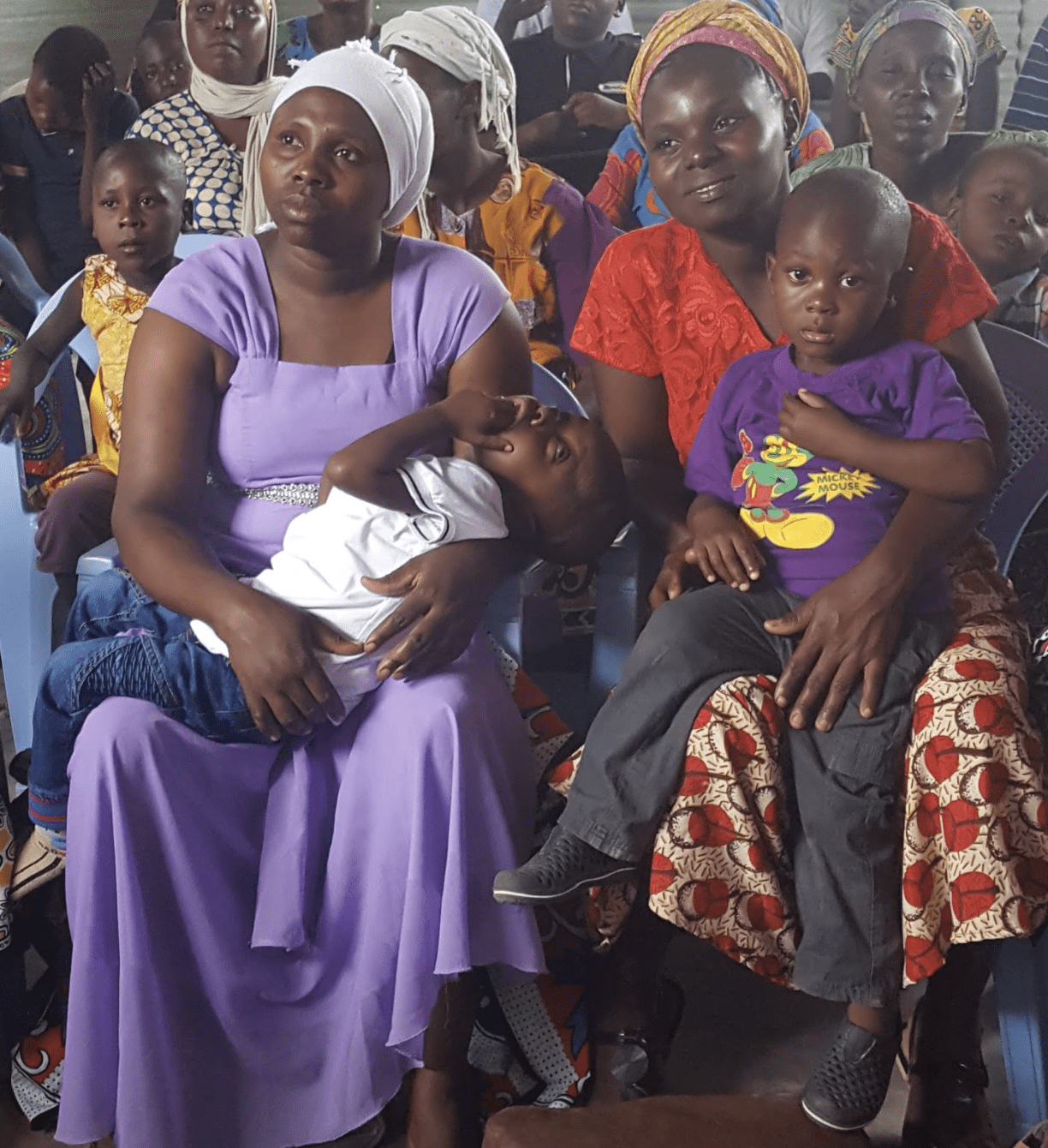 A group of Kenyan mothers, holding their children and listening to a speaker.