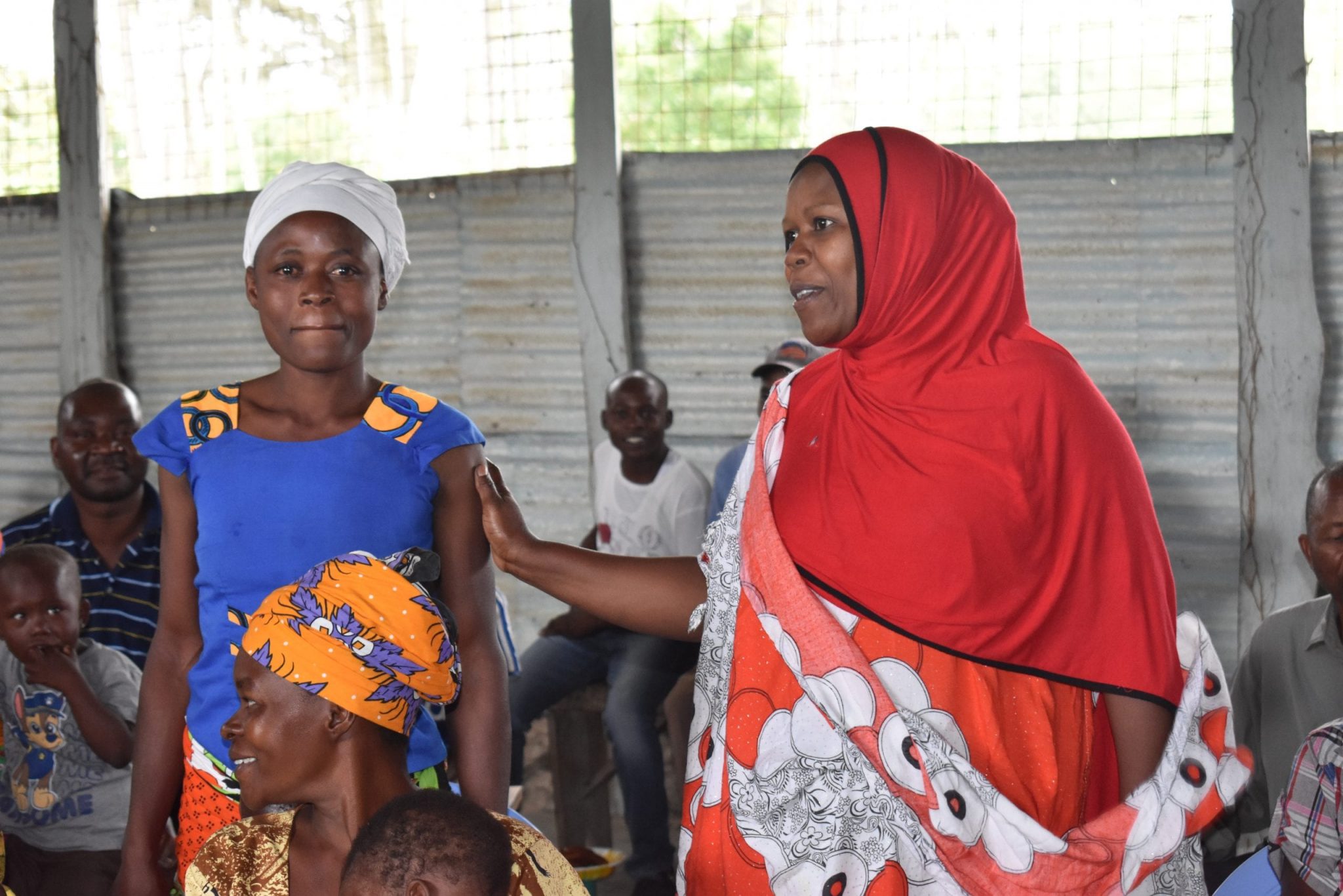 Two Kenyan women standing up with a group of others seated around them.