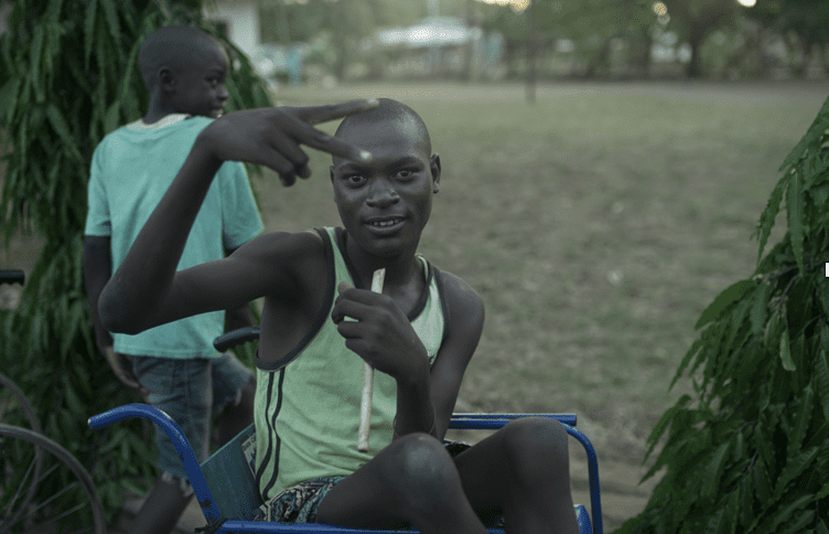 Boy wearing green tank top, sitting in blue wheelchair, making a "peace sign" with his hand.