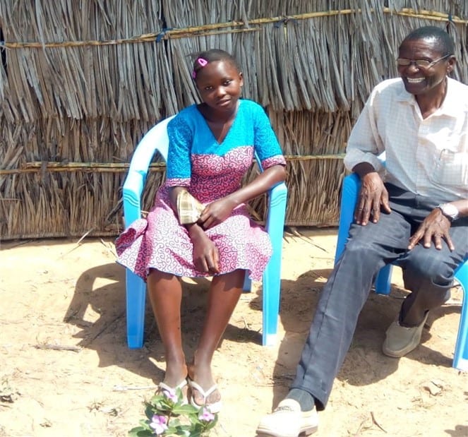 Kenyan pastor sitting beside a young girl in blue chairs outside