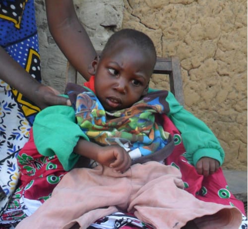 A young Kenyan child seated on a wooden chair, receiving help to stay sitting up.