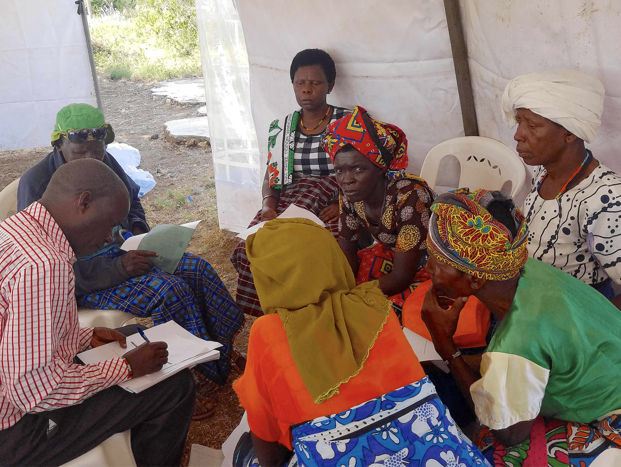 A group of Kenyan adults sitting inside a tent, holding pens and paper