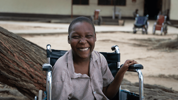A girl smiling and seated outside in a wheelchair, wearing a brown checkered shirt.
