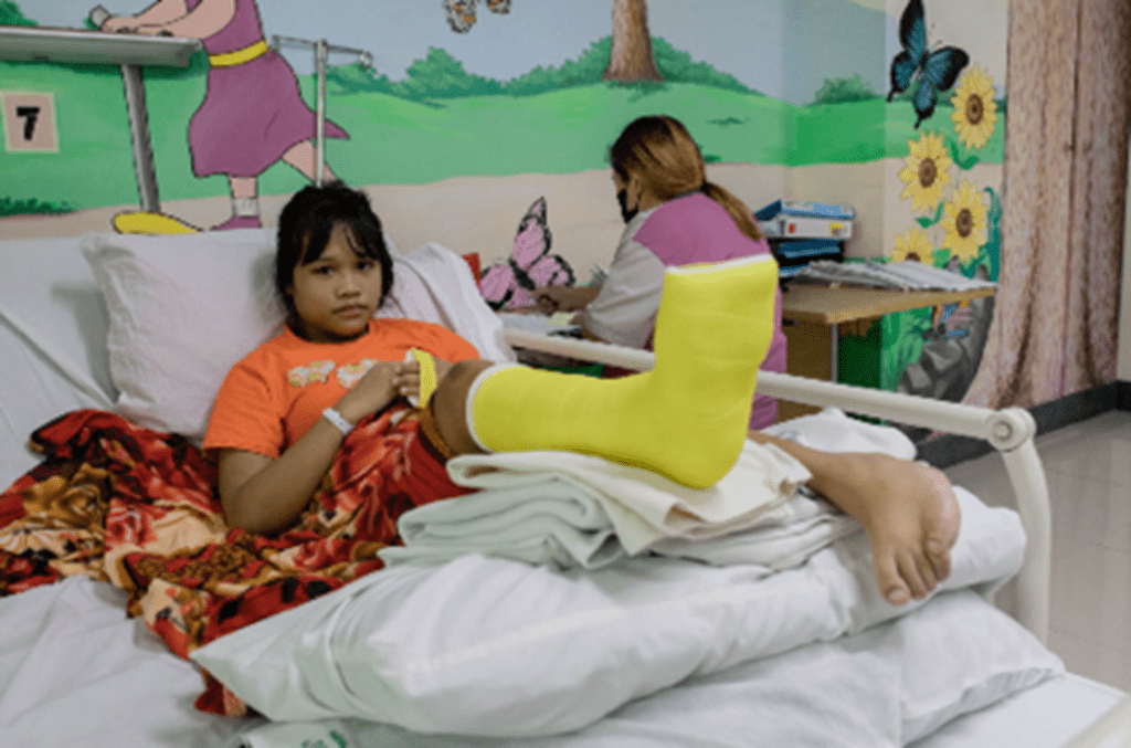 A young girl pictured in a hospital bed in a yellow cast and orange shirt