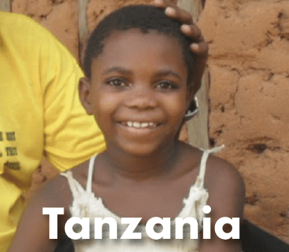 A smiling young girl with short hair wearing a white tank top, standing in front of an orange mud wall writing Tanzania on the image
