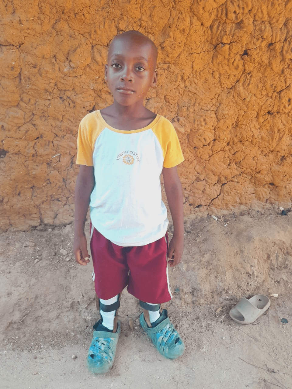 A boy in a yellow and red outfit standing against an orange mud wall, looking directly at the camera.