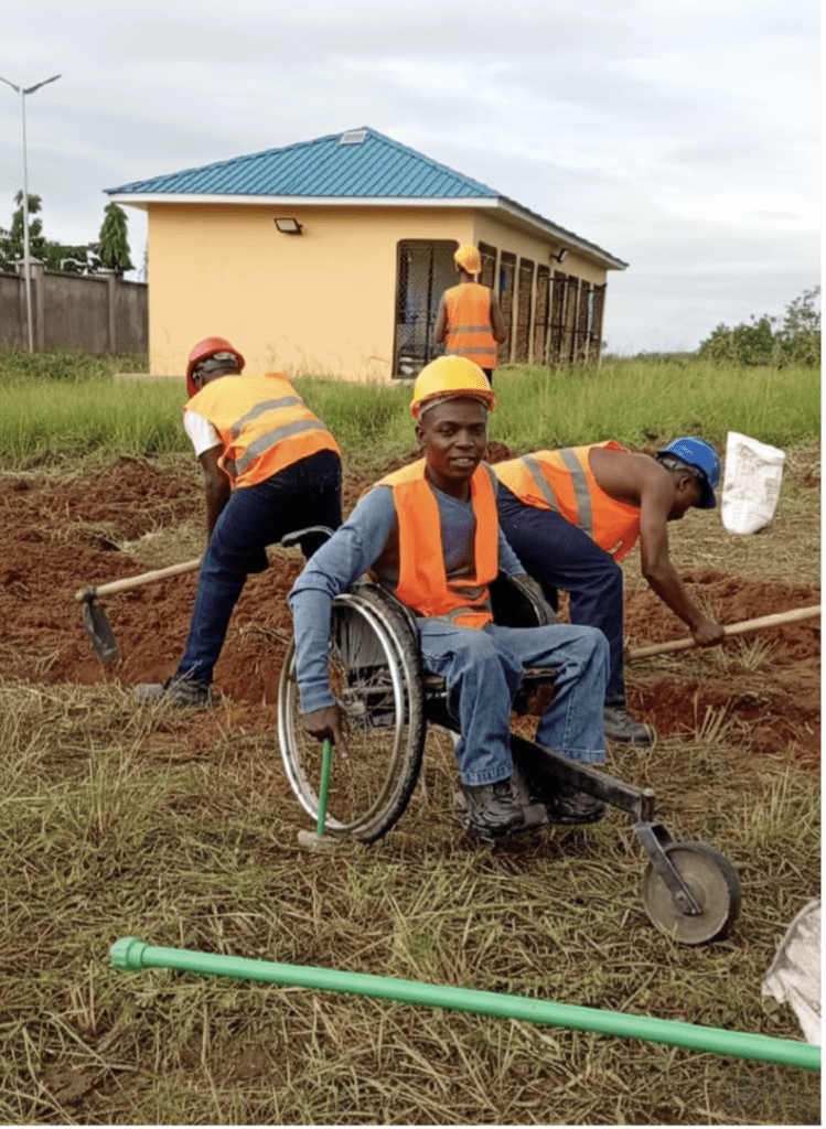 A person using a wheelchair works alongside others wearing safety vests and hard hats at a construction site, with teammates digging and installing materials near a small building in the background.