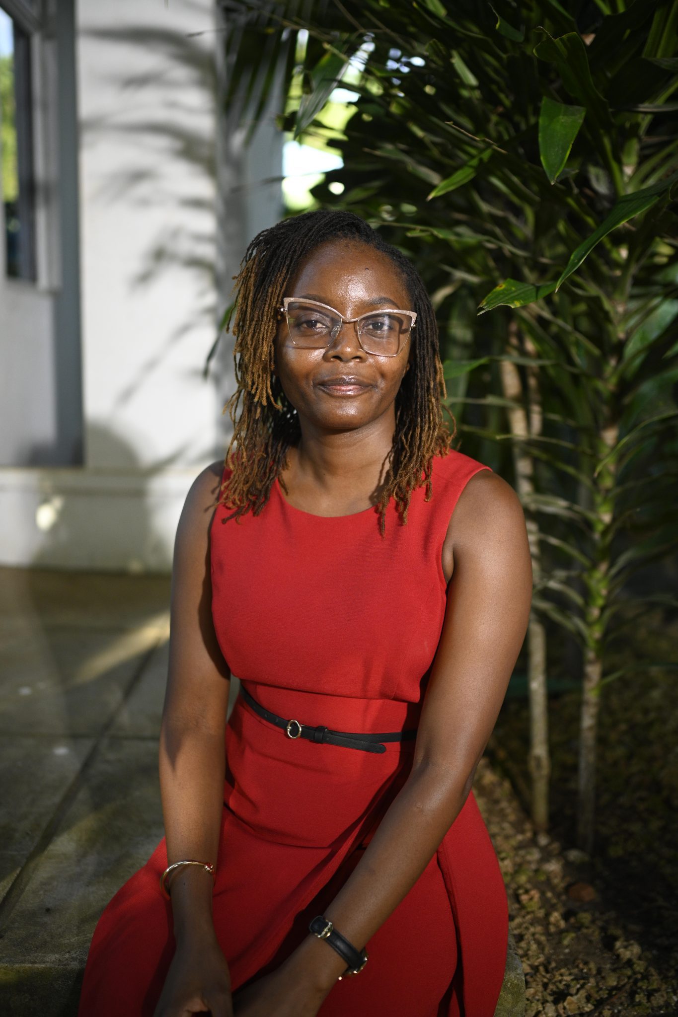 A Kenyan woman wearing glasses and a red dress, seated outside and posed for a photo.