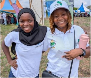 A young woman wearing a white shirt and white hat, and a teenage girl wearing a black hijab and white shirt. They are standing together outside and smiling for the camera.