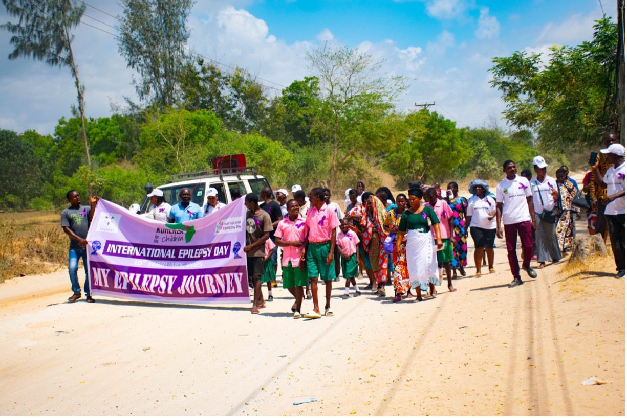 A group of children and adults walk together along a rural road carrying a large purple banner that reads “International Epilepsy Day – My Epilepsy Journey,” participating in a community awareness march in Kenya.