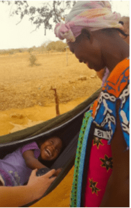 A woman wearing a colorful headscarf smiles down at her laughing child resting in a hammock in an outdoor rural setting