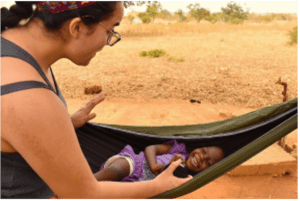 A woman wearing glasses smiles and reaches toward a laughing child lying in a hammock outdoors in a rural setting.