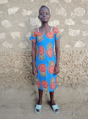 A young girl stands against a textured, weathered wall, wearing a bright blue dress with red circular patterns and light-colored sandals. She faces the camera with a calm, neutral expression, her hands resting at her sides.