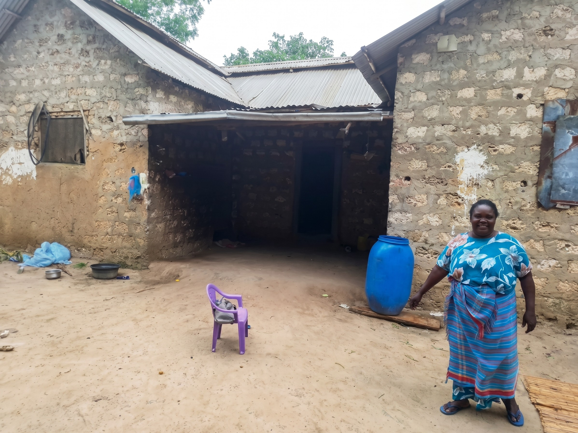 Woman standing outside a stone home with a blue water barrel and a small child’s chair in the courtyard.