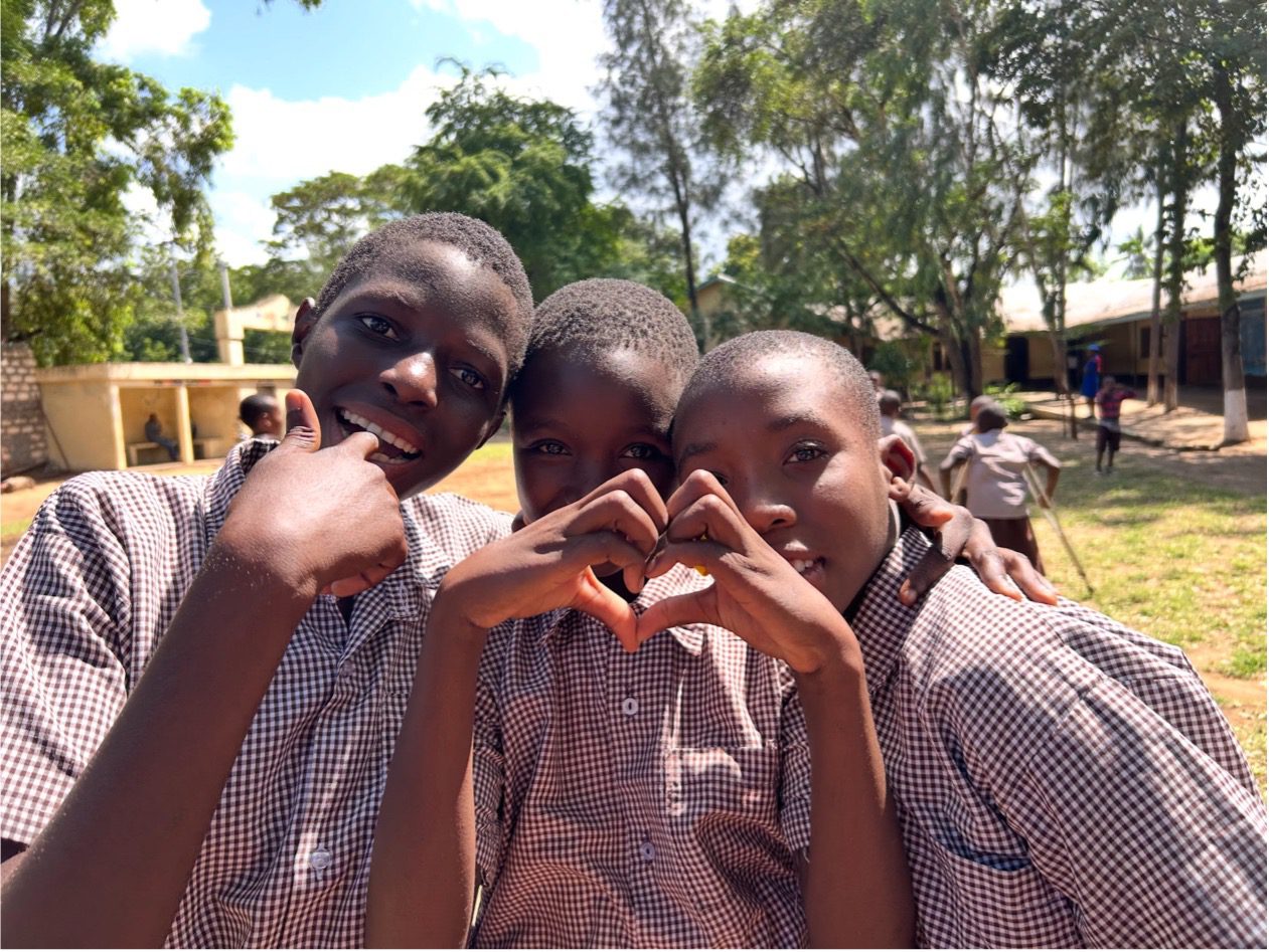 Three smiling students in matching school uniforms pose together outdoors, with one student forming a heart shape with their hands