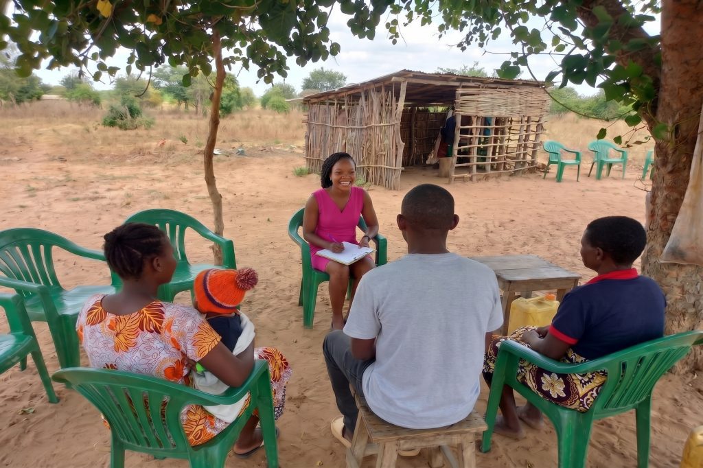 A family sitting outside in rural Kenya, being counseled by a woman wearing a pink dress