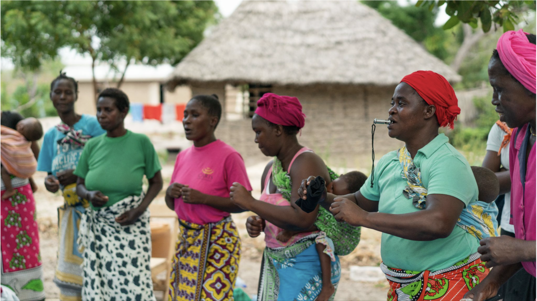 A group of African women gathered outdoors in a rural village, wearing colorful clothing and patterned wraps. Several women carry children, while a woman in a green shirt and red headscarf plays a small wind instrument. A thatched-roof building and trees are in the background.